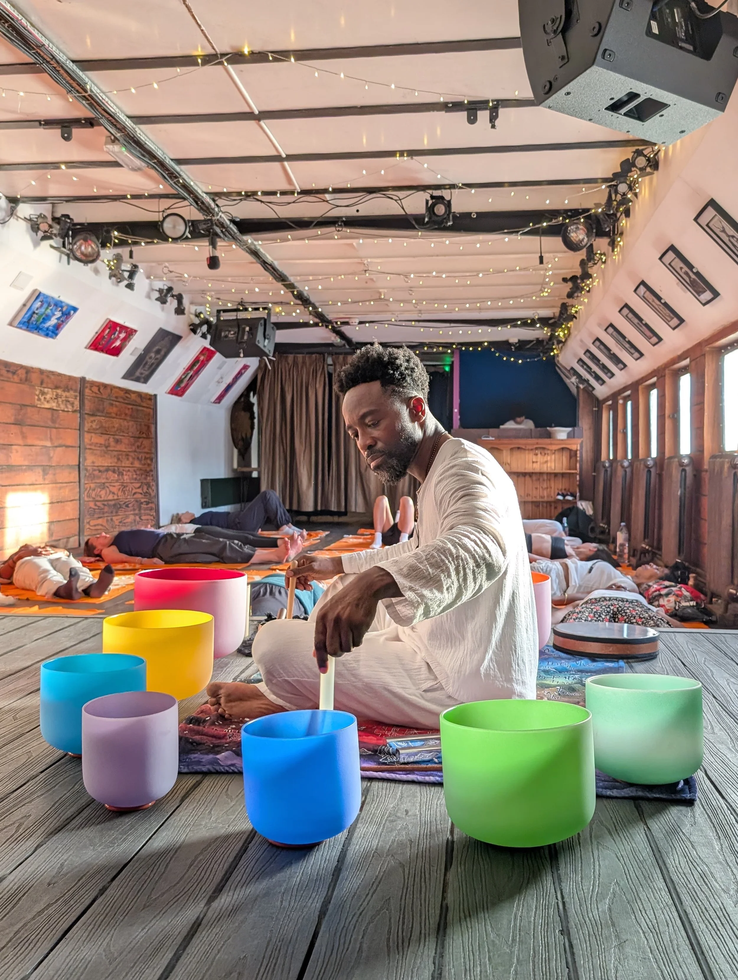 A group of people participating in a meditation or yoga session indoors, with some lying down and one person sitting cross-legged surrounded by colorful singing bowls, with string lights on the ceiling and framed pictures on the walls.