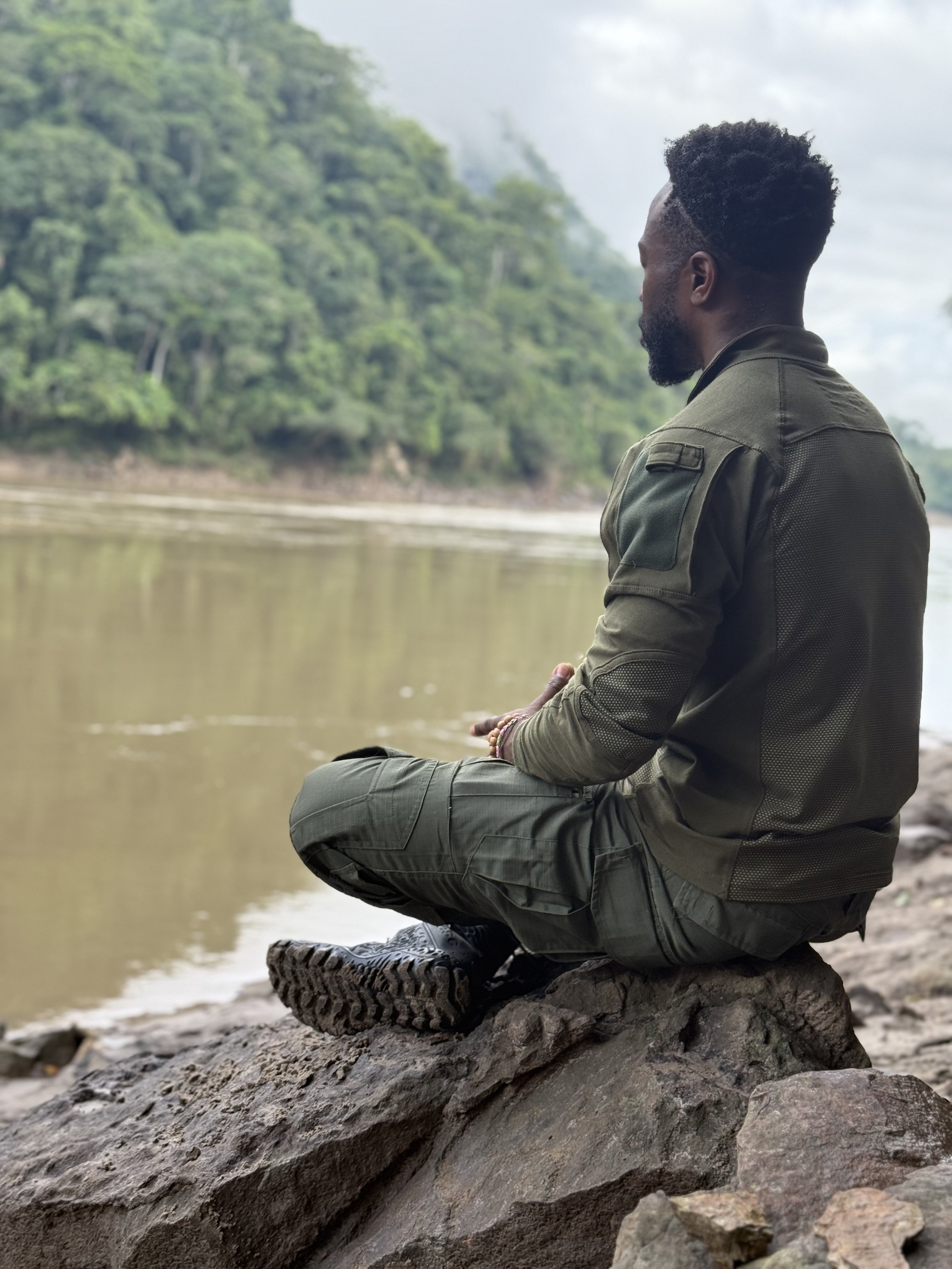 A man in outdoor clothing sitting cross-legged on a rock by a body of water, with trees and cloudy sky in the background.