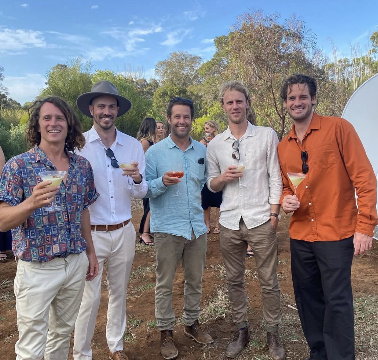 A group of five men standing outdoors at a gathering, holding drinks and smiling at the camera, with other people and trees in the background.
