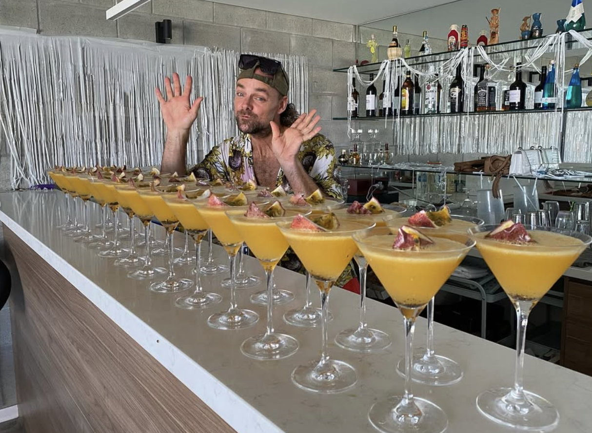 A man with a beard and floral shirt, wearing sunglasses and a cap, behind a bar with multiple yellow cocktails garnished with fruit slices. The bar is decorated with white streamers and bottles of alcohol on shelves.