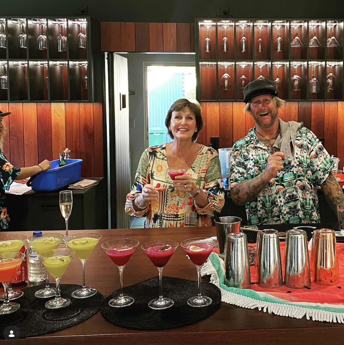 Two people smiling and holding colorful cocktails at a bar with various drinks and glasses on the counter.