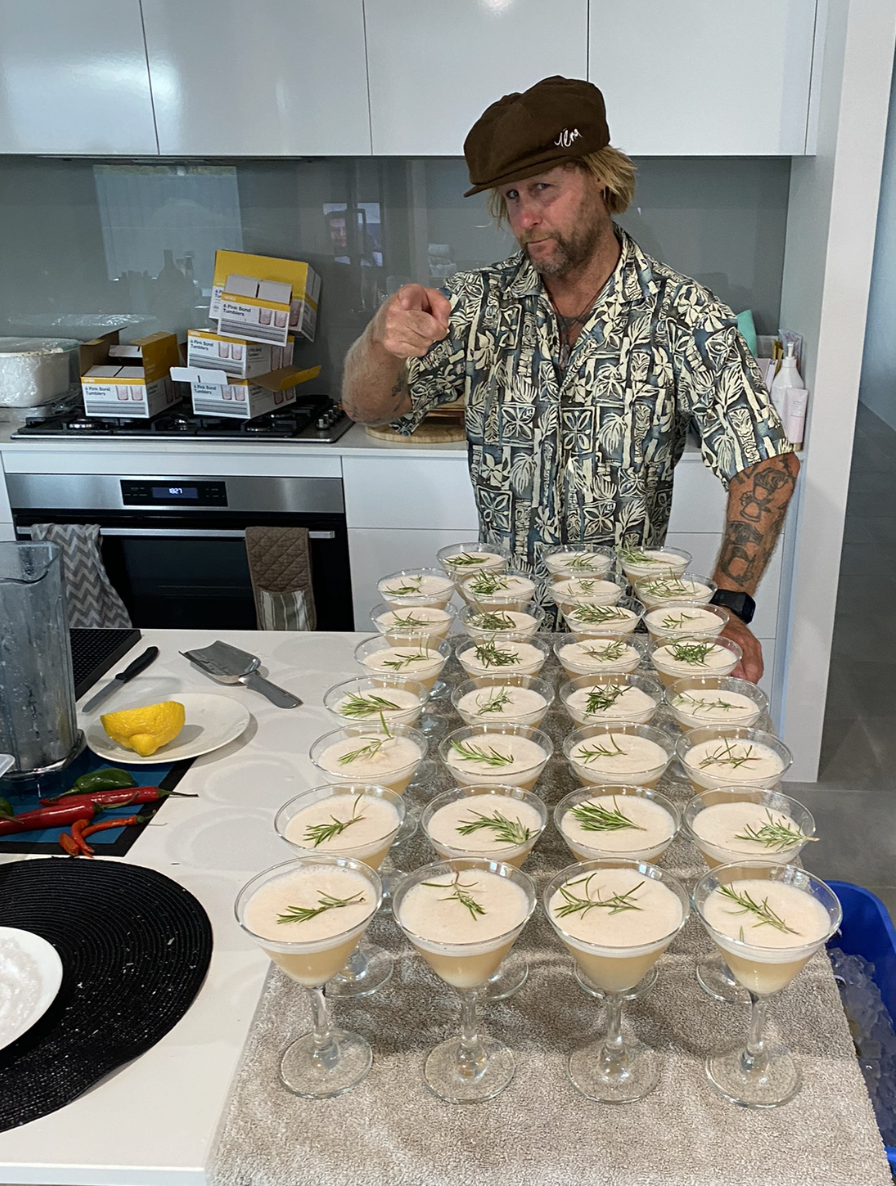 A man in a Hawaiian shirt and a brown cap pointing at the camera, standing behind a table with multiple glasses of a creamy beverage garnished with rosemary sprigs, in a modern kitchen.