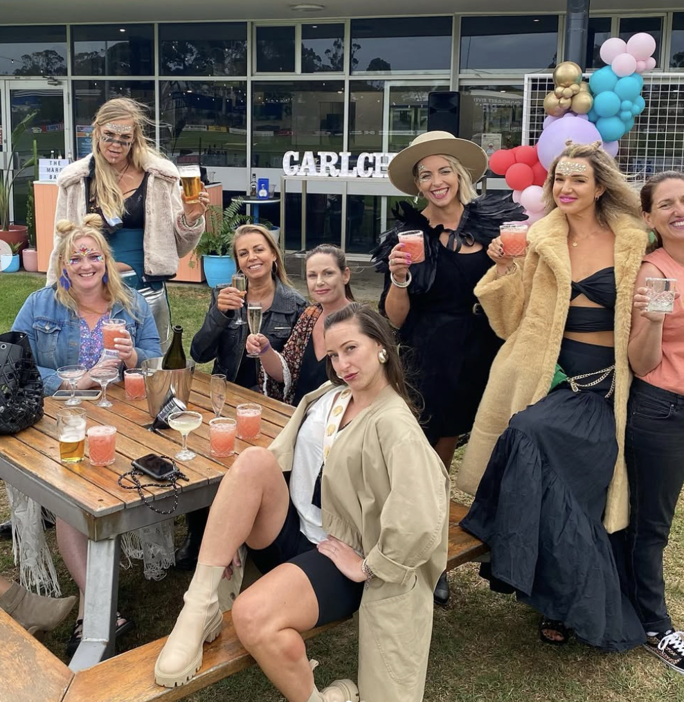 Group of women celebrating outdoors with drinks in front of a building decorated with balloons and a sign that says 'GARDEN'.