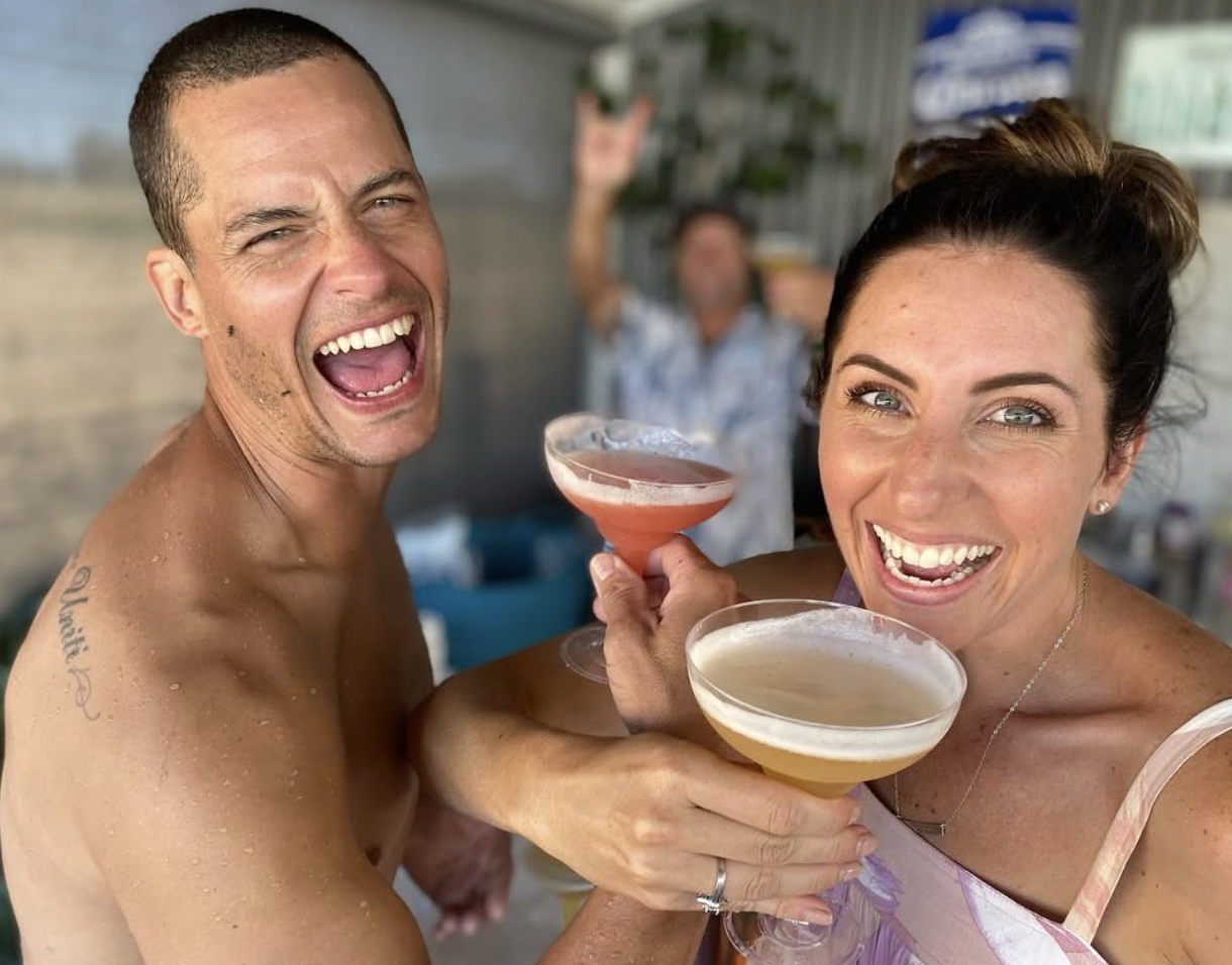 A man and woman smiling and holding cocktails at a social gathering, with two blurred people in the background.