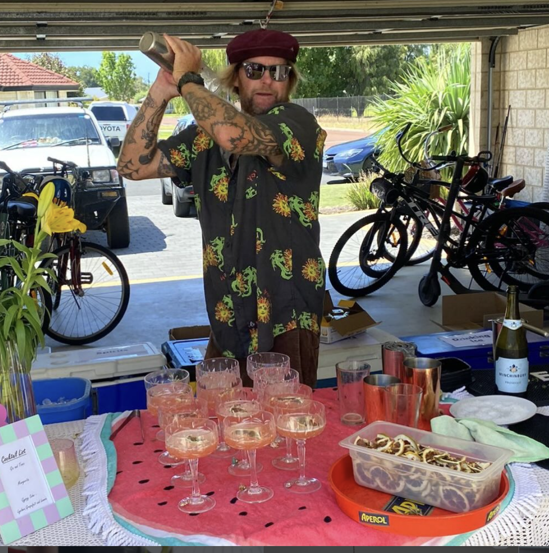 A man with tattoos, wearing sunglasses and a colorful short-sleeve shirt, holding a cocktail shaker behind his head in a garage or outdoor patio area. There are several drinks on a table in front of him, along with snacks, a bottle of champagne, and bicycles in the background.