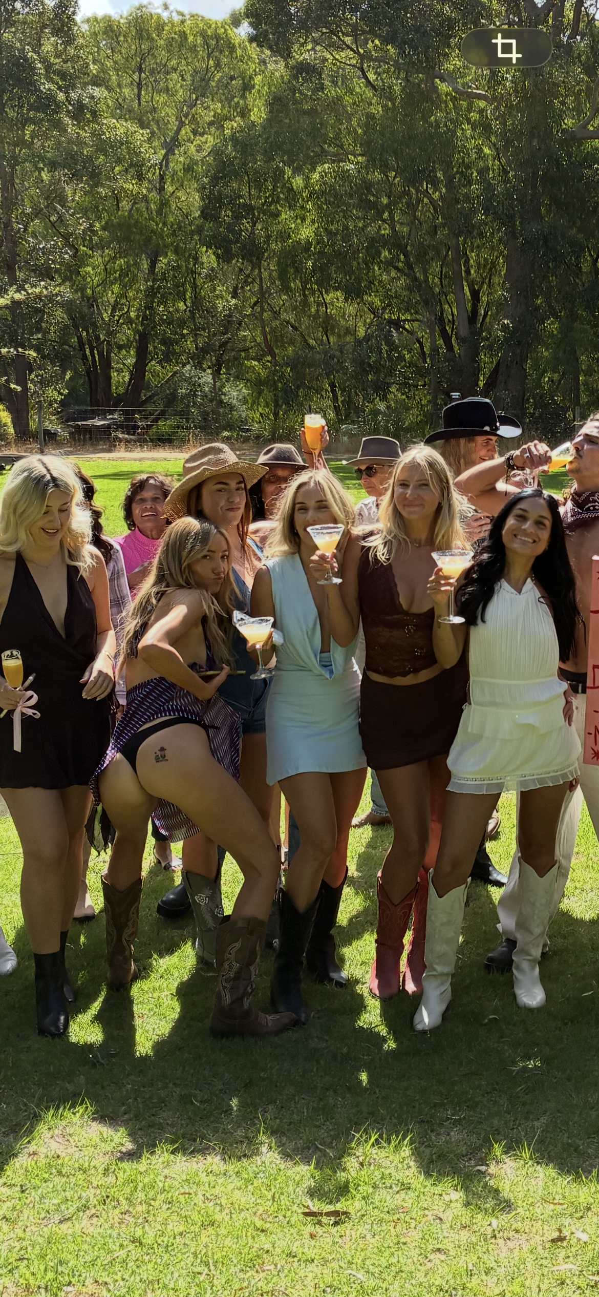 Group of women outdoors at a celebration, holding cocktails, dressed in stylish and cowboy-themed clothing, with green trees and grass in the background.