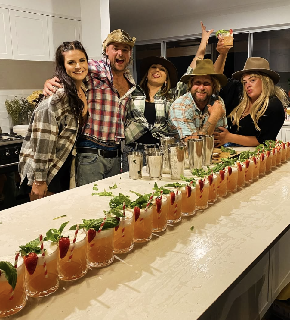 Group of five friends in plaid and cowboy hats celebrating with a line of cocktails garnished with strawberries and mint on a kitchen counter.