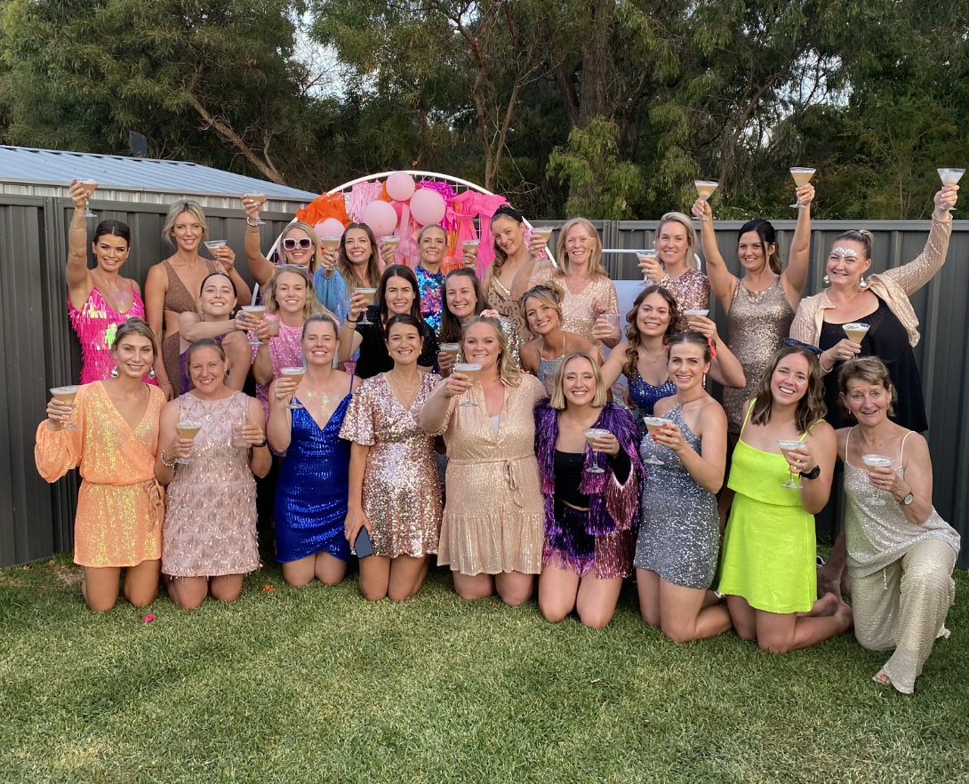 Group of women celebrating outdoors at a party, dressed in colorful dresses, holding up cocktails and smiling, with balloons and pink decorations in the background.
