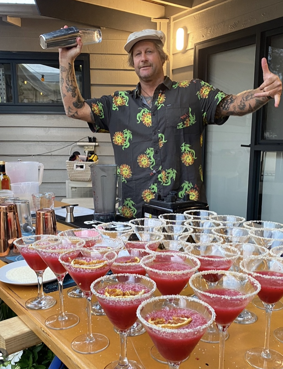 A man with tattoos wearing a white cap and a colorful pineapple and dragon shirt, holding a cocktail shaker and making a gesture with his other hand, standing behind a table filled with pink cocktails with salted rims and fruit garnishes, outdoors on a porch.