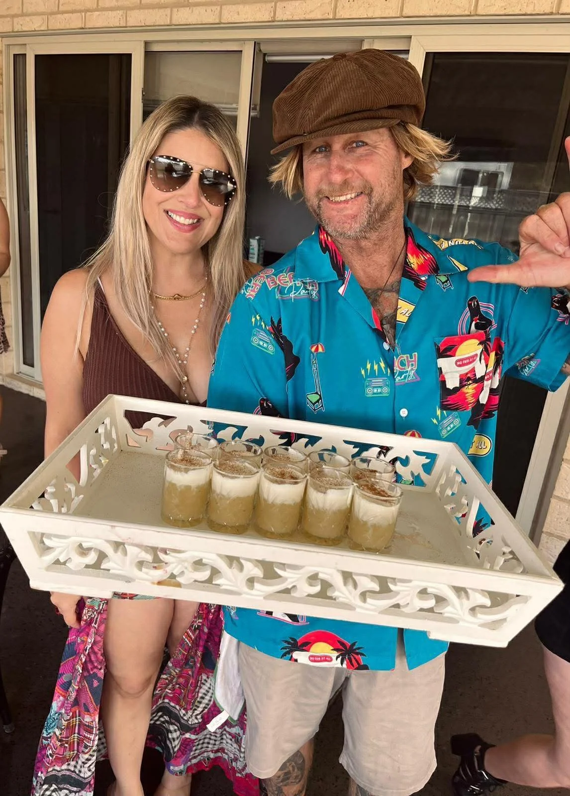 A man and woman smiling, holding a tray with multiple glasses of a layered dessert. The woman is wearing sunglasses and a sleeveless top, and the man is wearing a colorful tropical shirt and a brown cap. They are standing outside near a sliding glass door.