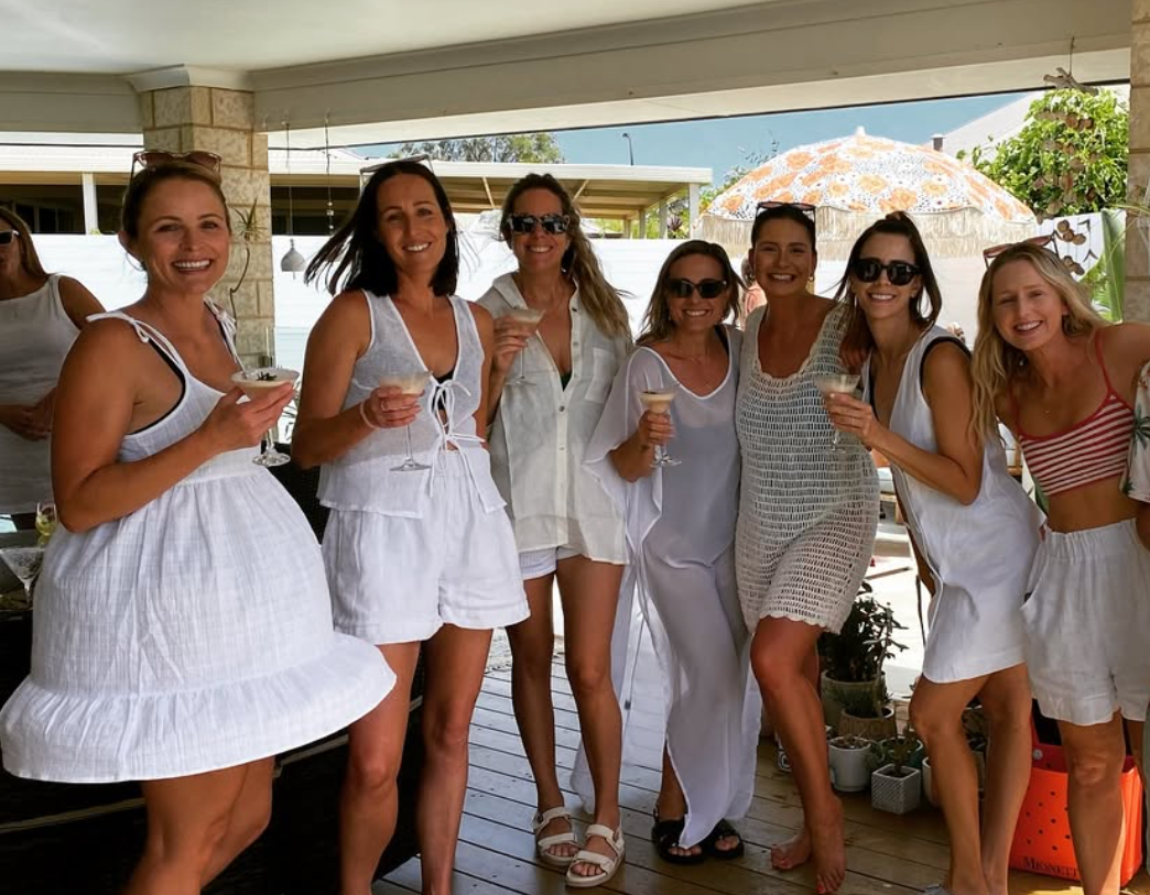 Group of women in summer attire celebrating with drinks on a covered patio, some holding glasses, with a large umbrella and potted plants in the background.