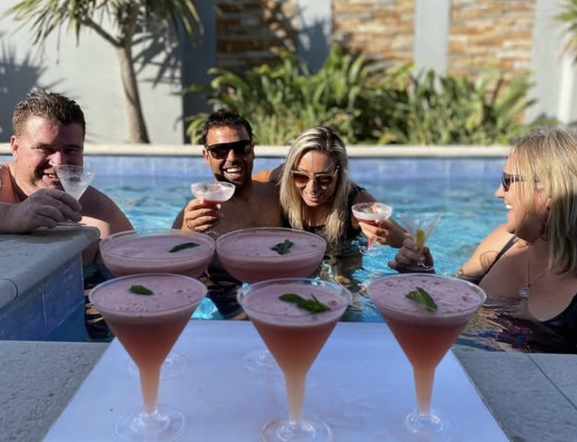 Group of four friends enjoying drinks in a pool, with pink cocktails in the foreground.