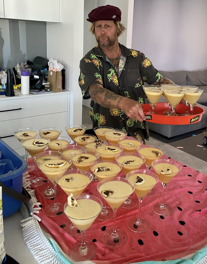 A man with blonde hair and a beard wearing a colorful Hawaiian shirt and a maroon hat holding a red tray with drinks, standing behind a table filled with many yellow cocktails garnished with fruit, on a red watermelon-patterned cloth.