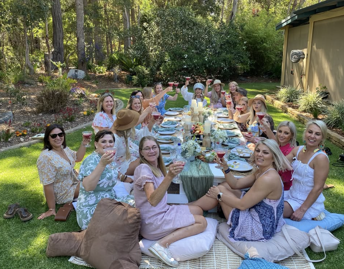A group of women enjoying a picnic outdoors, sitting around a long table with food and drinks, raising their glasses in a toast, on a sunny day in a lush garden.