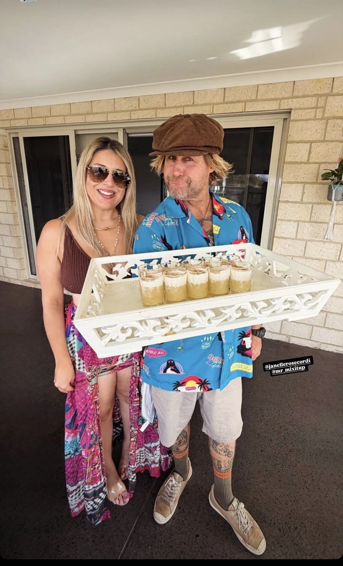 A man and woman posing outdoors, the man holding a tray of seven glasses filled with drinks topped with whipped cream. The woman is wearing sunglasses and a sleeveless top, while the man is wearing a colorful Hawaiian shirt, shorts, and a brown cap. They are standing on a patio with a brick wall in the background.