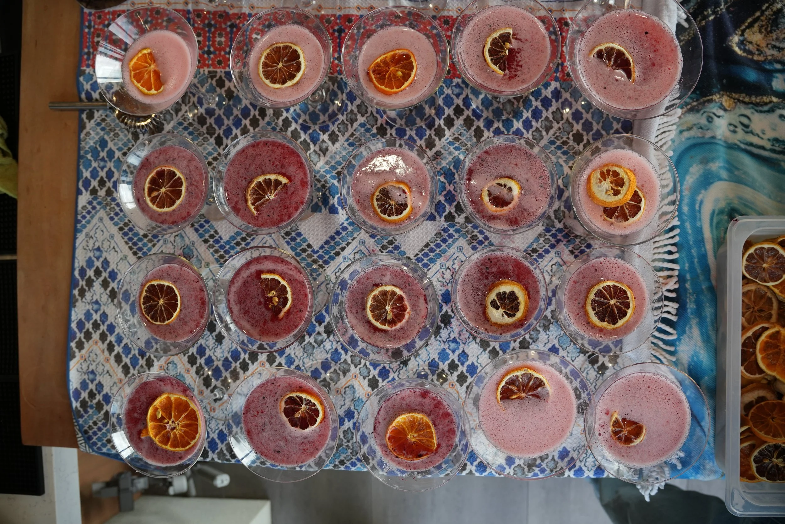 Multiple glasses of pink beverage garnished with dried citrus slices arranged on a patterned tablecloth.