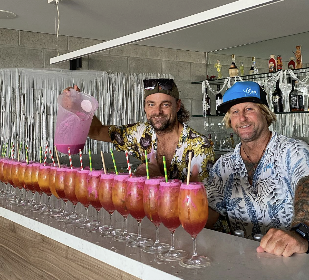 Two men celebrating at a bar with a row of colorful drinks, the man on the left pouring a pink drink into the glasses and the man on the right smiling, wearing a blue baseball cap.