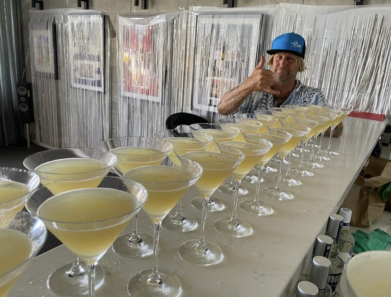 Man wearing a blue hat and tropical shirt giving a thumbs up behind a long countertop with many yellow cocktails in martini glasses.