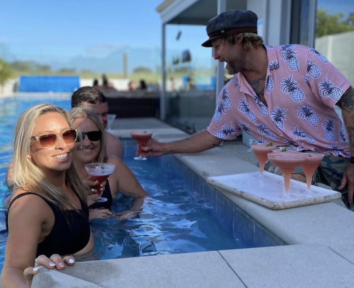 People enjoying drinks at a pool party, with a man in a pineapple-patterned shirt serving pink cocktails to women wearing swimsuits and sunglasses.