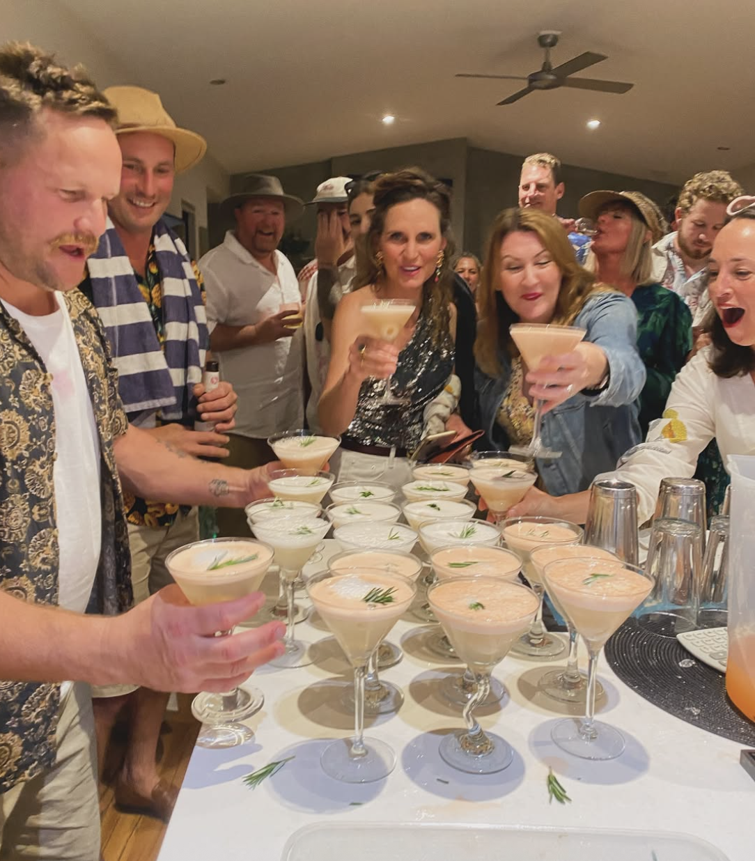 Group of people celebrating with cocktails at a party, standing around a table with many drinks garnished with herbs.