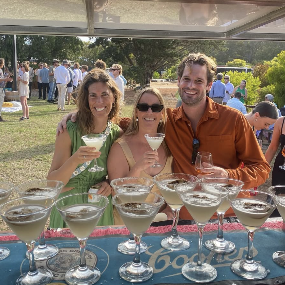 Three people smiling and holding cocktails at an outdoor gathering with a table full of drinks in the foreground.