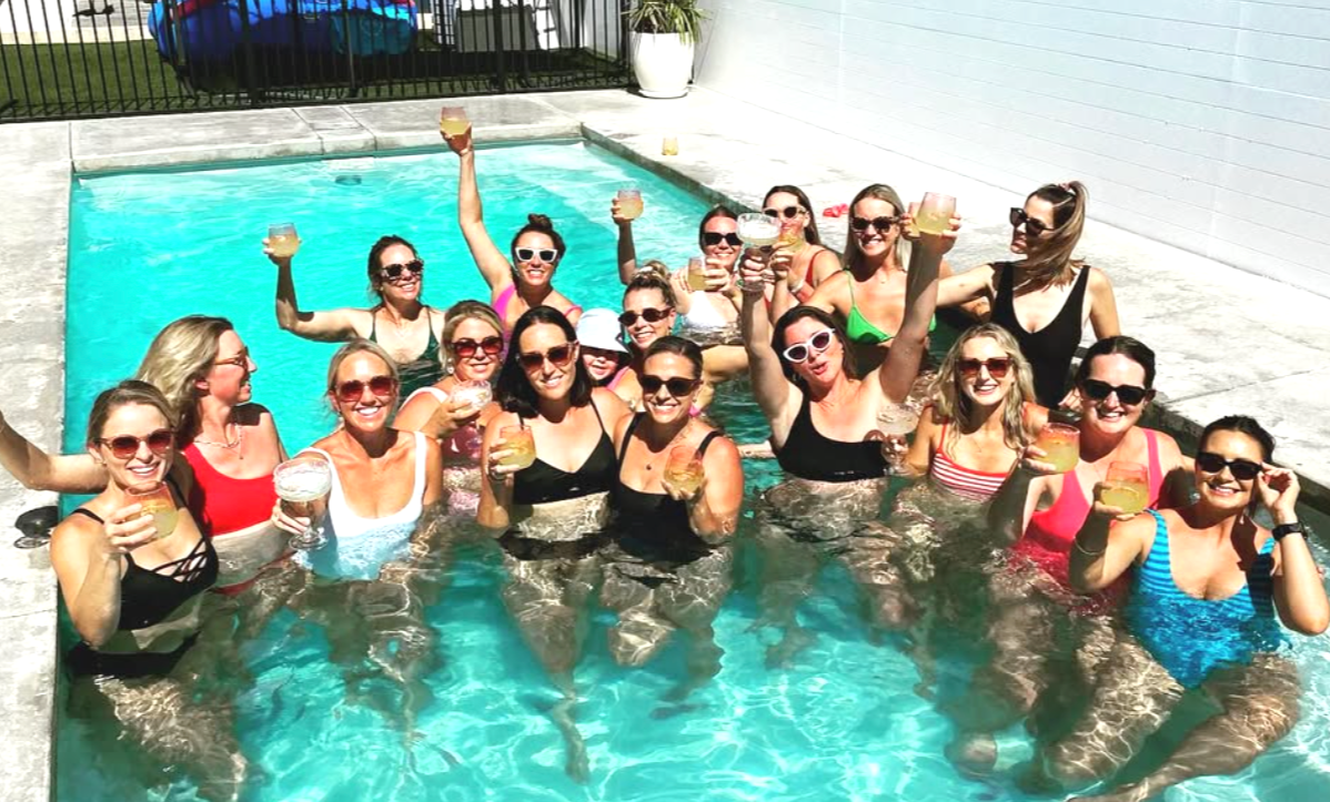 Group of women in swimsuits and sunglasses enjoying a poolside gathering, holding drinks and celebrating.