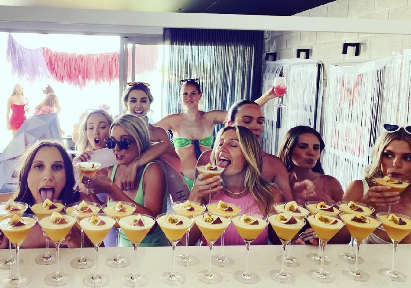 A group of women at a celebration, possibly a bridal shower, enjoying drinks and surrounded by yellow desserts topped with fruit on a counter.