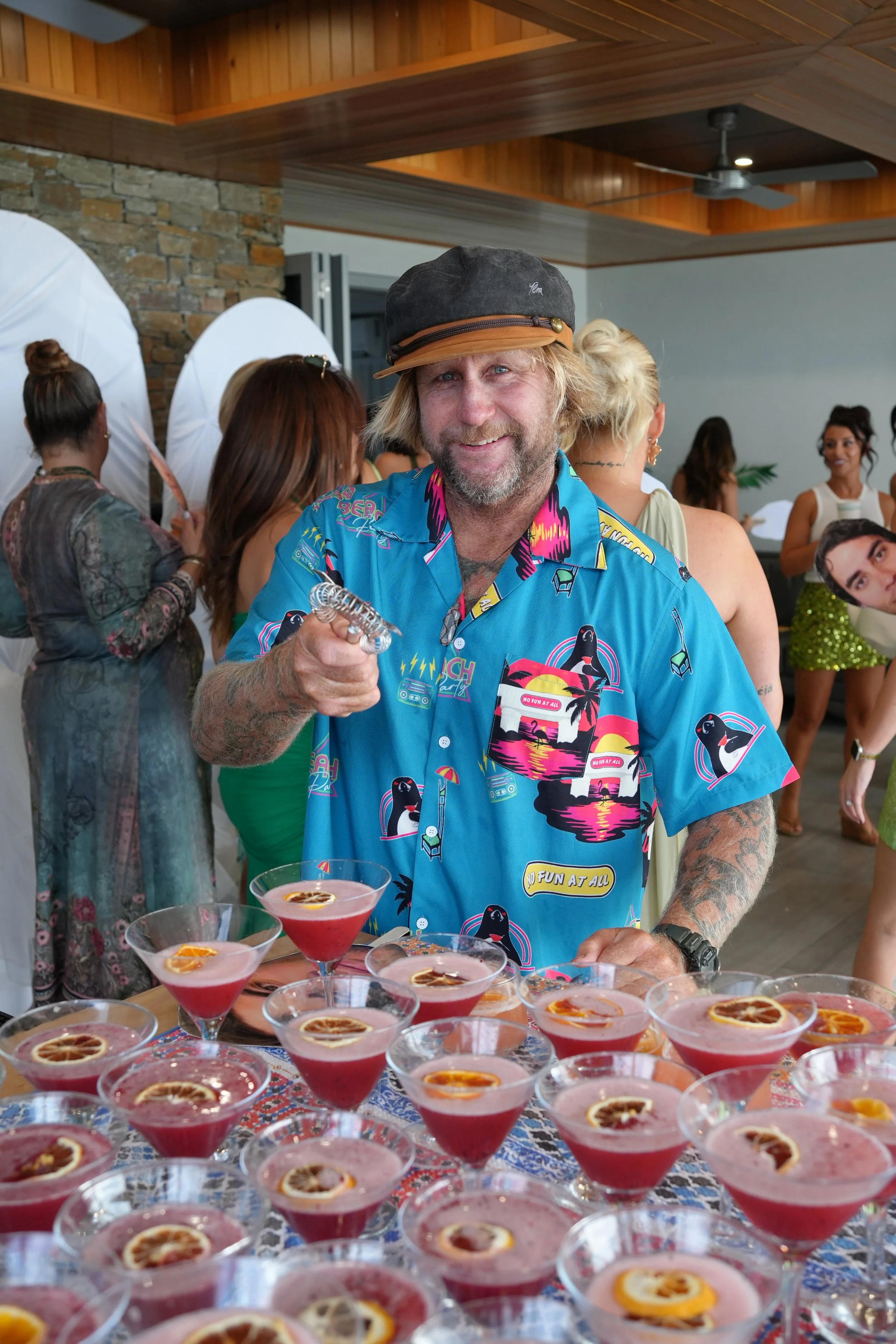 A man with a beard, blond hair, and a colorful Hawaiian shirt stands behind a table of red and pink cocktails garnished with orange slices, in a room with wooden ceiling, brick wall, and group of people in the background.