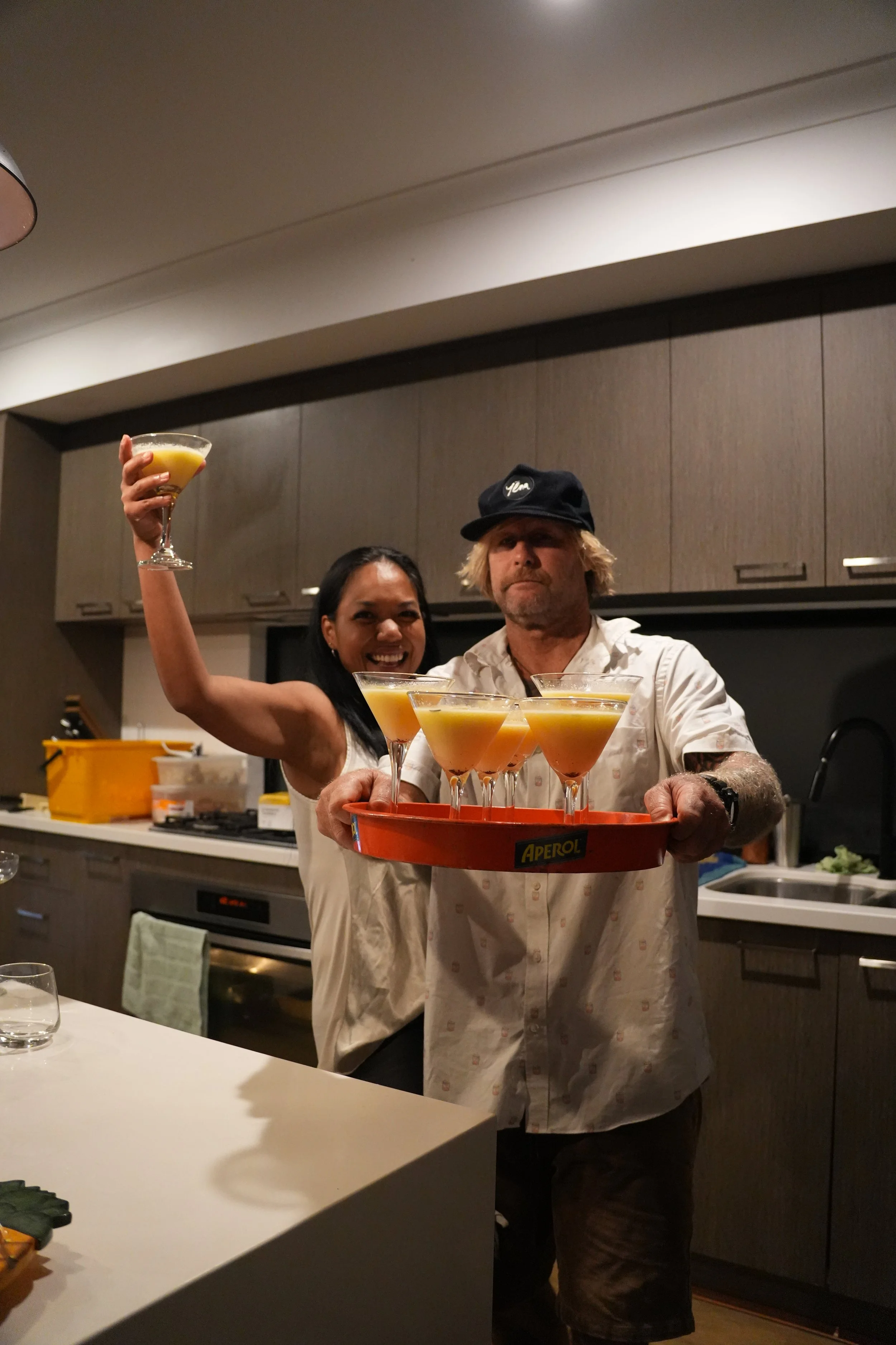 A woman and a man in a kitchen holding a tray of yellow cocktails. The woman is smiling and holding a cocktail glass in her right hand, while the man is holding the tray with multiple cocktails. The kitchen has cabinets, a stove, and various kitchen items in the background.