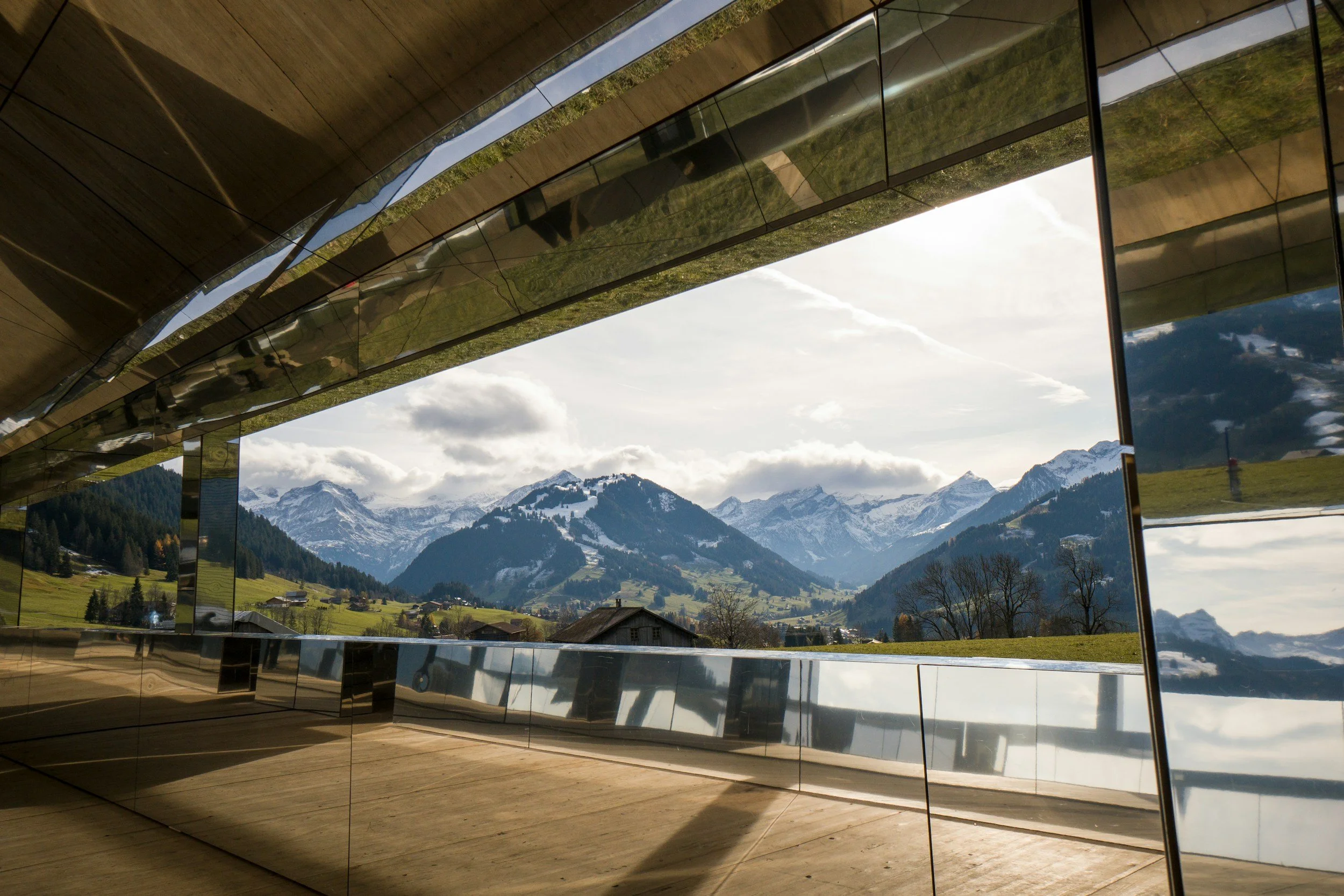 Vue d'un bâtiment moderne avec des murs en miroir reflétant un paysage de montagnes enneigées et un ciel nuageux.