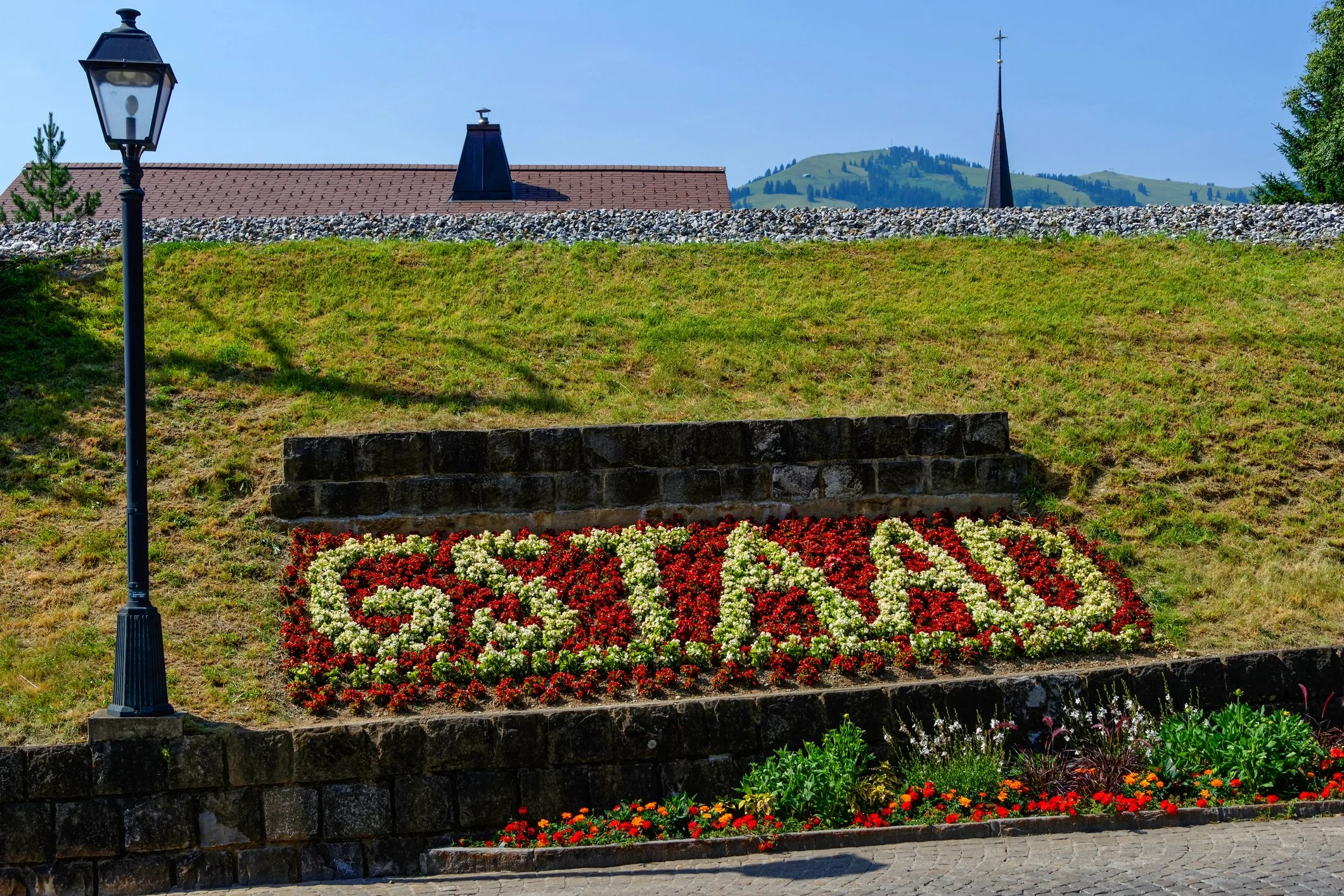 Jardin avec des fleurs rouges, blanches et oranges, une pente d'herbe verte, un lampadaire noir à gauche, un mur en pierre, un ciel bleu clair, un toit en tuiles rouges, une tour d'église avec une croix, montagnes en arrière-plan.