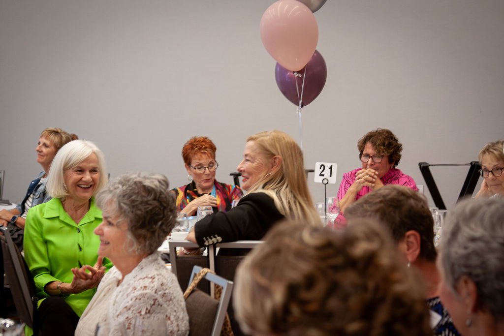 A group of elderly women sitting at tables during a social event, with some laughing and others observing.