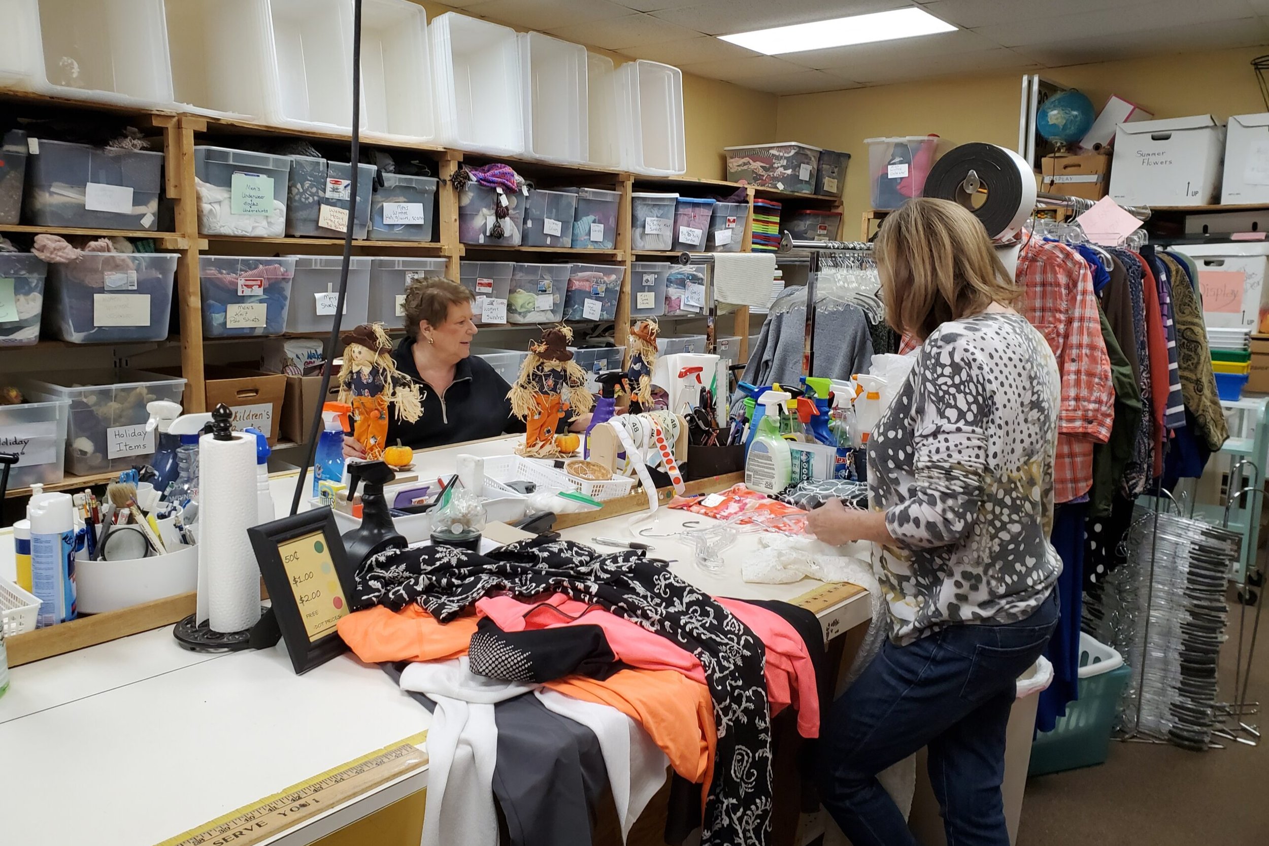Two Thrift Shop volunteers pricing donations for resale. Shelves in the background are filled with plastic bins containing various items labeled for organization.