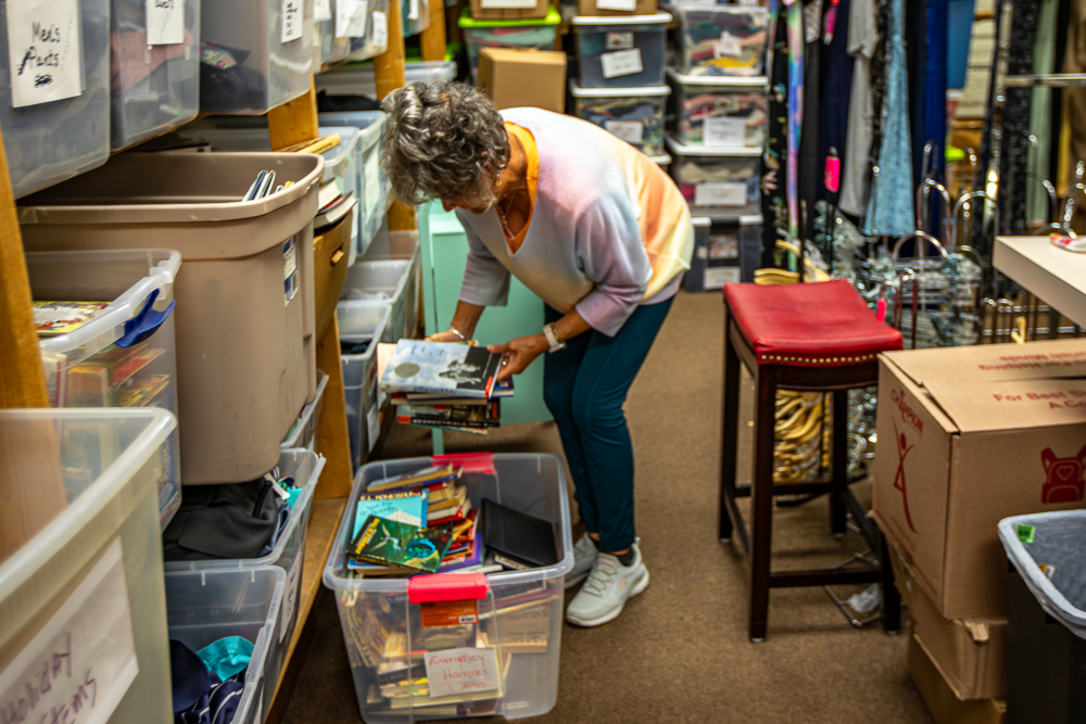 A volunteer is looking through a plastic bin filled with books in the Thrift Shop storage room with shelves of supplies and clothing in the background.