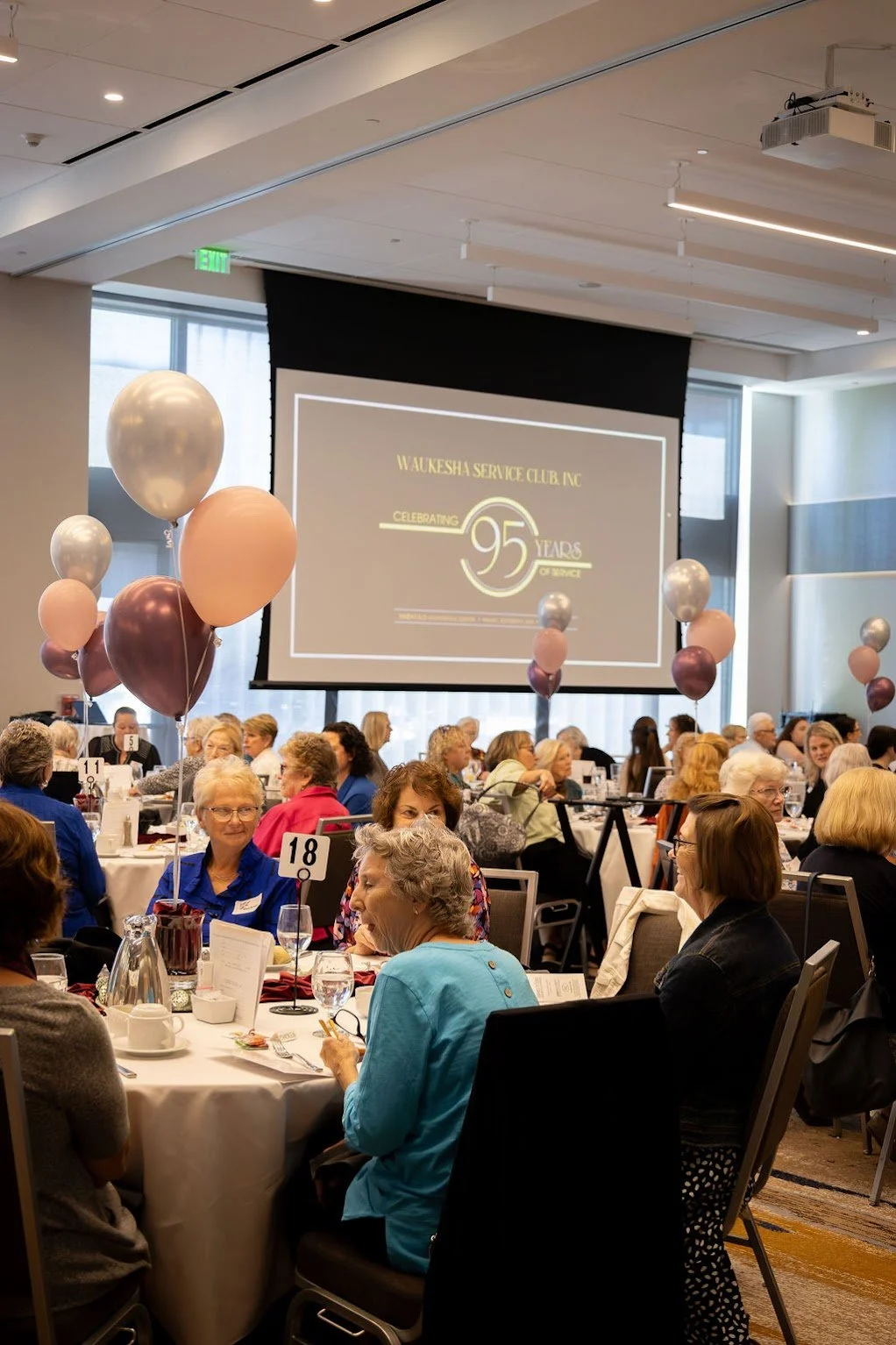 Women attending a banquet celebrating the 95th anniversary of Waukesha Service Club, Inc., with balloons, round tables, and a large screen displaying event information.