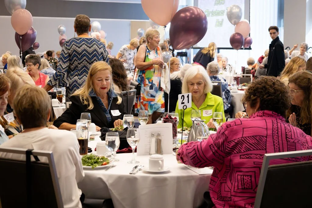 A large group of laughing and smiling women gathered around a banquet table decorated with white tablecloths, glassware, and floral centerpieces.