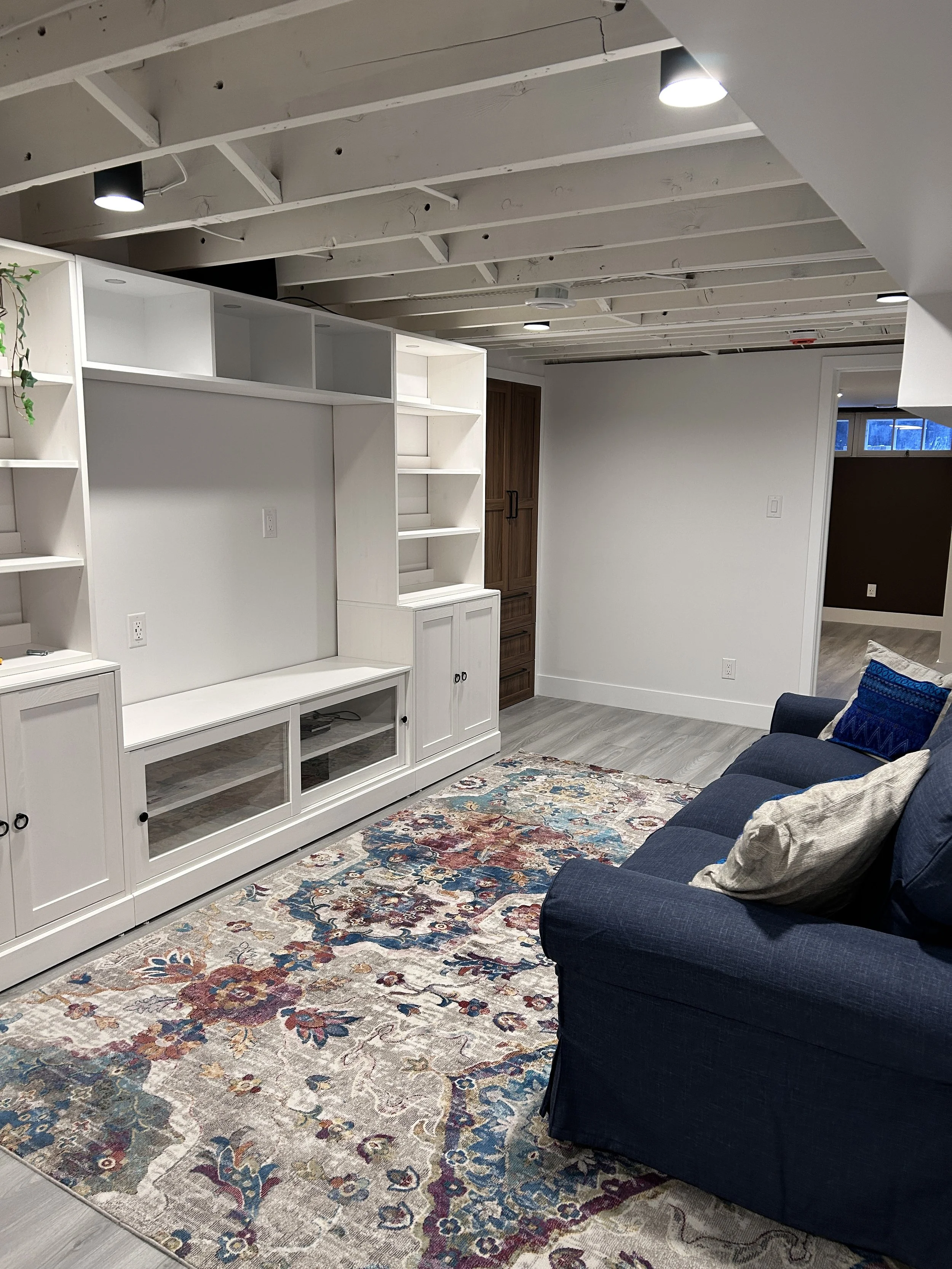 Living room with a white entertainment center, a blue sofa with pillows, a colorful rug, and an exposed ceiling with lights. In Winnipeg Manitoba
