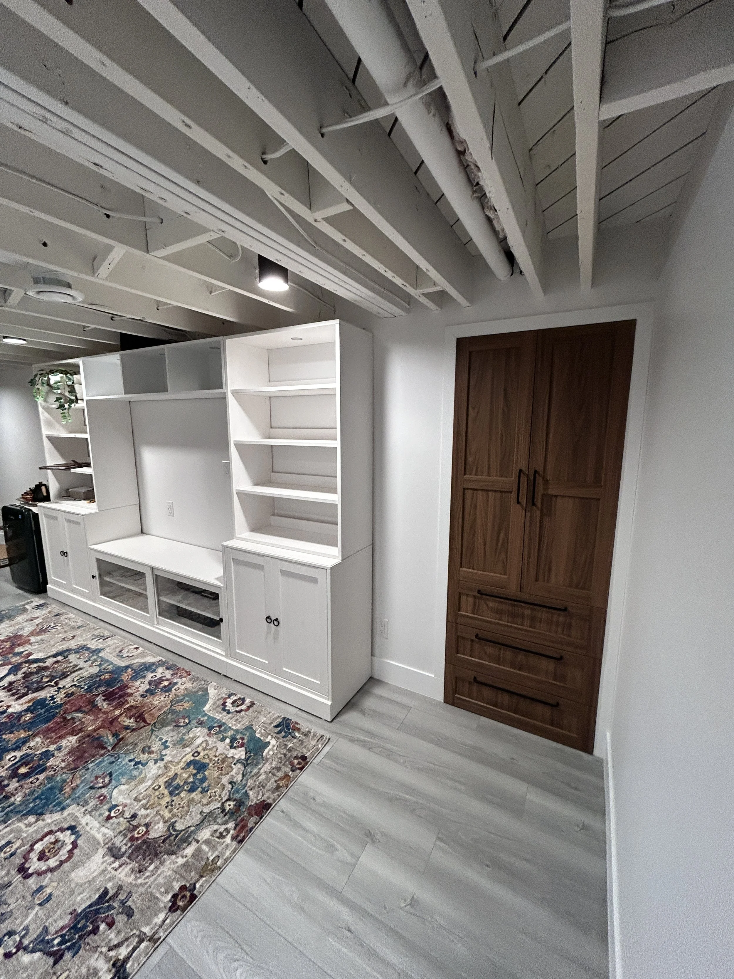 Interior space showing white built-in entertainment center with open shelving and cabinets, a wooden cabinet with black handles, a multicolored area rug, and a ceiling with exposed beams and pipes. Winnipeg