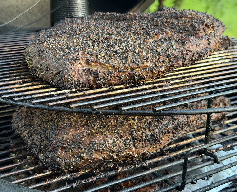 Two seasoned, pepper-rubbed briskets slow-smoking over charcoal on a grill.