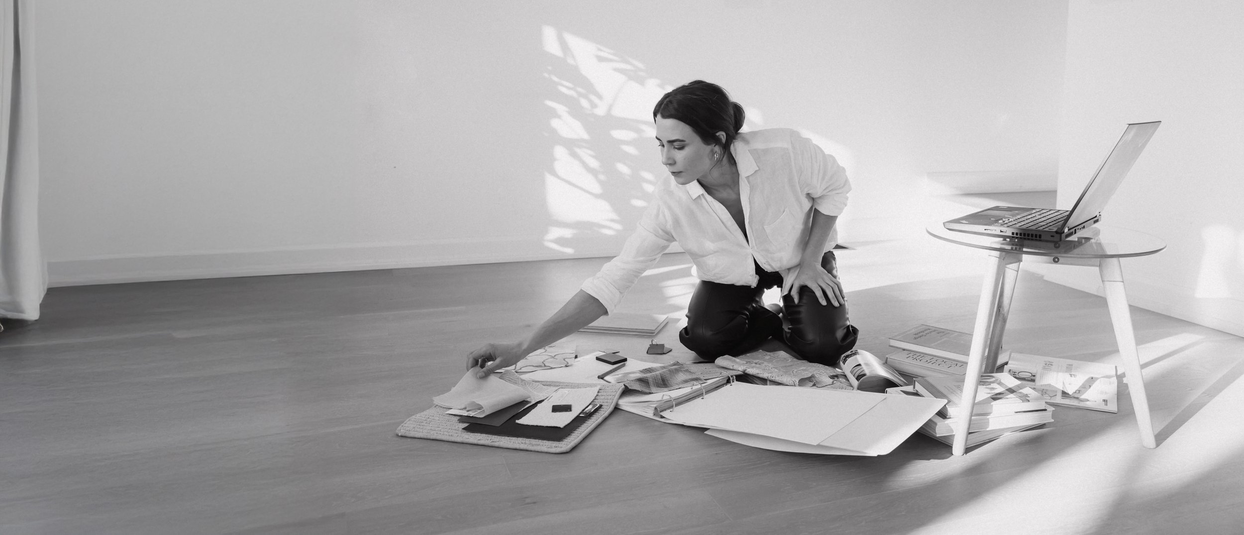 A woman in a white shirt is kneeling on the floor, sorting through magazines and papers, with a laptop on a small table nearby, in a bright room with wooden flooring.
