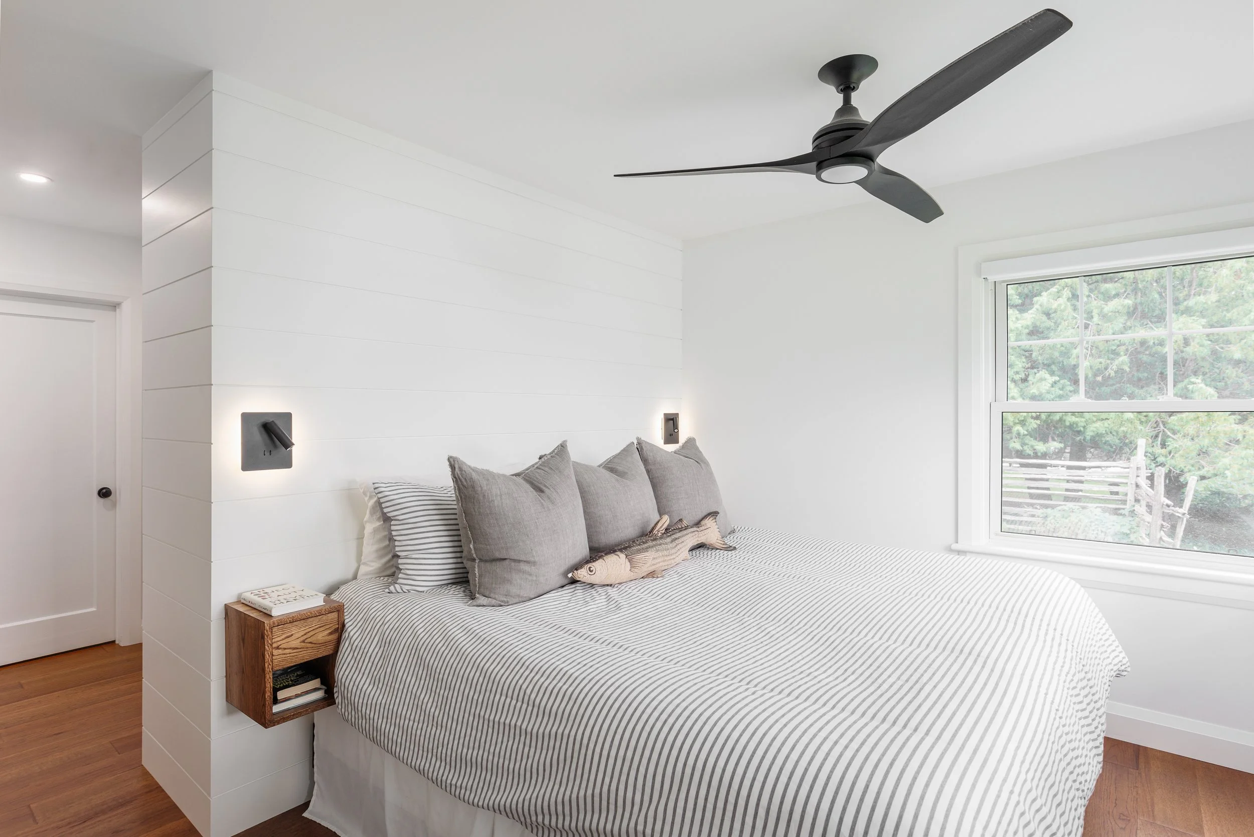 A bedroom with a white wall, a large window showing greenery outside, a bed with striped bedding and multiple gray pillows, a wooden bedside table with books, and a ceiling fan.