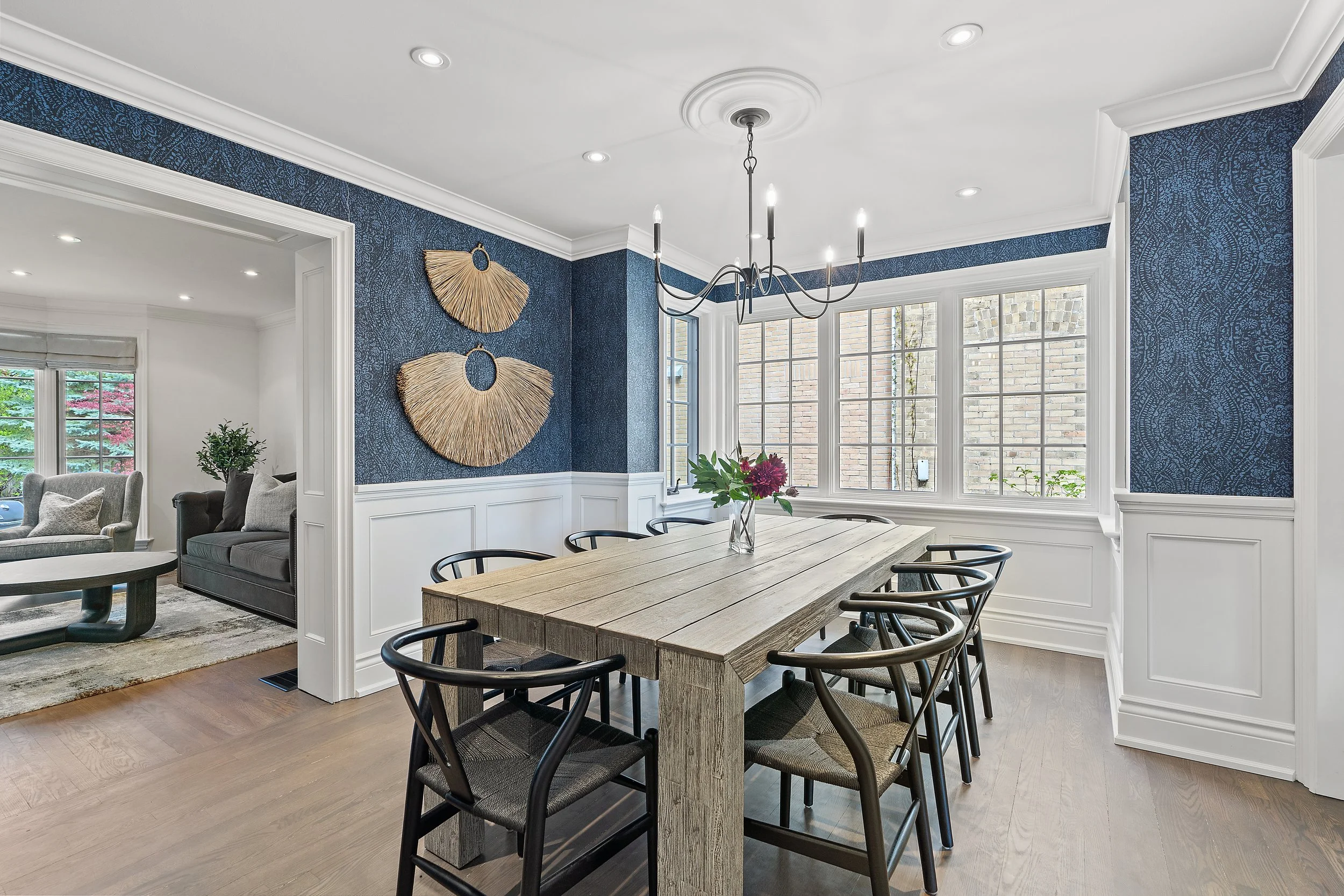 A dining area with a wooden table surrounded by black chairs, a chandelier hanging from a white ceiling, large windows, and a blue textured wall with woven wall art.