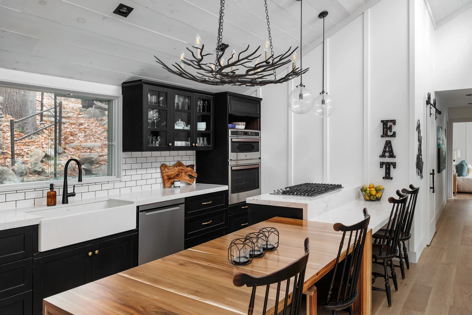 A modern kitchen with black cabinets, white countertops, a white farmhouse sink, a large window showing an outdoor scene with leaves and rocks, a chandelier with a branch-like design, pendant lights, and a black and white color scheme.