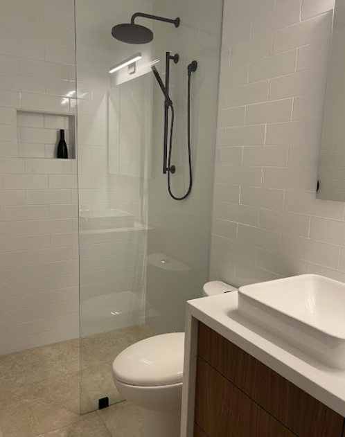 A modern bathroom with a glass-enclosed shower, a wall-mounted showerhead, a black soap bottle in a niche, a white toilet, a white rectangular vessel sink on a wooden vanity, and a mirror on the wall.
