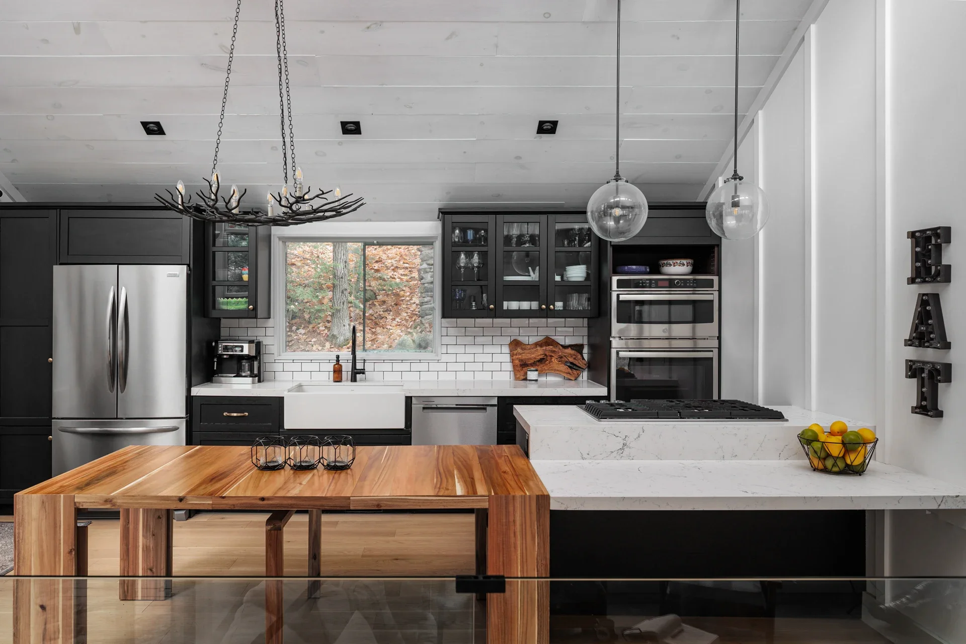 Modern kitchen with black cabinets, white subway tile backsplash, stainless steel appliances, wooden table, and pendant lights.