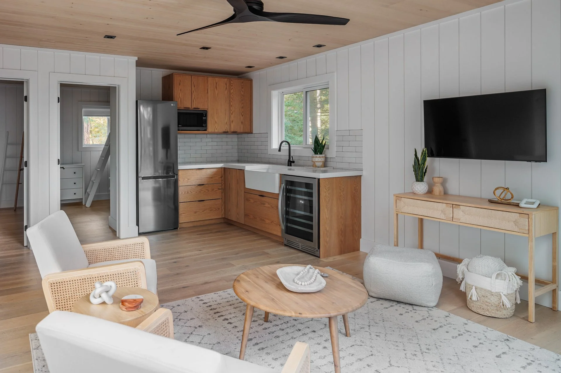 Living room area with a kitchen in the background, featuring a wooden ceiling fan, white paneled walls, a window with a view of trees, a wall-mounted TV, plants, a wooden table, and woven decor items.