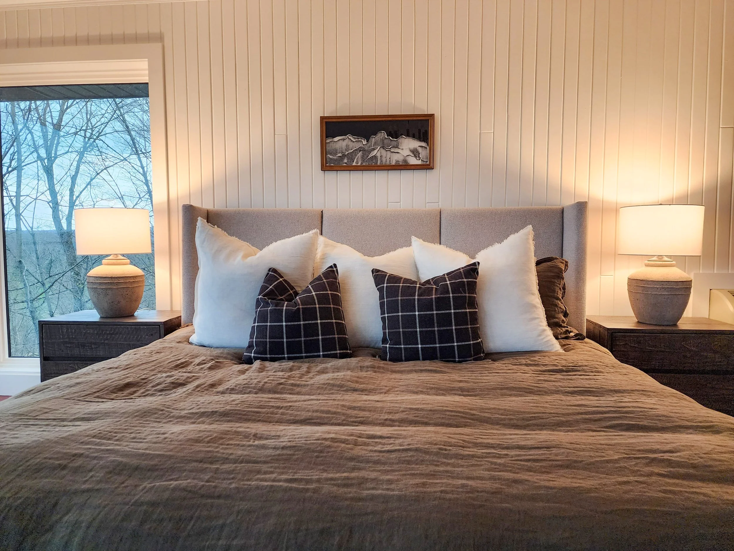 A bedroom with a large bed, decorative pillows, two nightstands with lamps, a framed mountain artwork above the bed, and a window showing leafless trees outside.