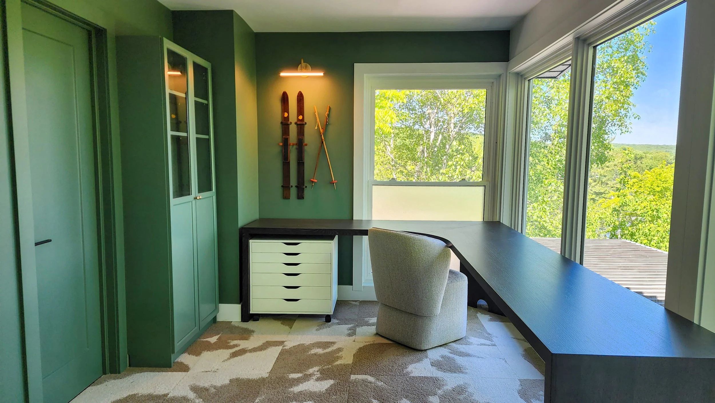 A home office with green walls, a large window showing trees outside, a black desk, a beige upholstered chair, a white rolling cabinet, a set of vintage skis mounted on the wall, and a small overhead light.