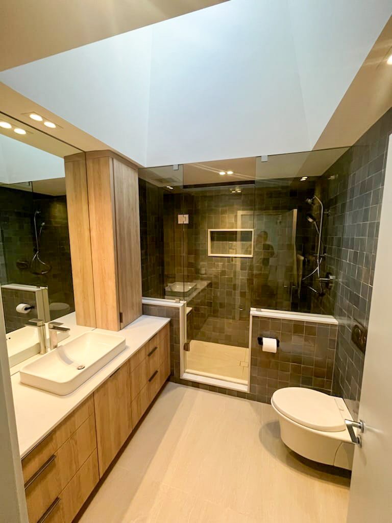 Modern bathroom featuring a wooden vanity with a vessel sink and a large mirror, a glass-enclosed shower with dark tiles, and a wall-mounted toilet.