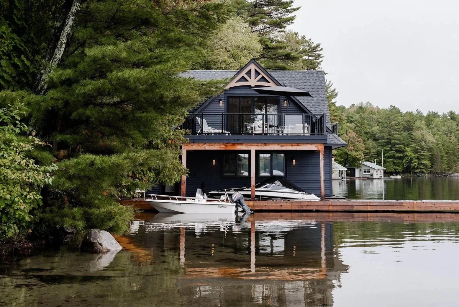 A two-story house with dark blue siding and a balcony on the upper level, situated by a lake surrounded by trees. There are boats docked at the house, and a reflection of the house can be seen in the water.